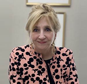 A woman with light hair in an updo, wearing a pink and black patterned blazer, smiles slightly, embodying the spirit of Douglas Marketing Group. The neutral background with framed artwork reflects the creative flair often found in a dynamic digital marketing company.