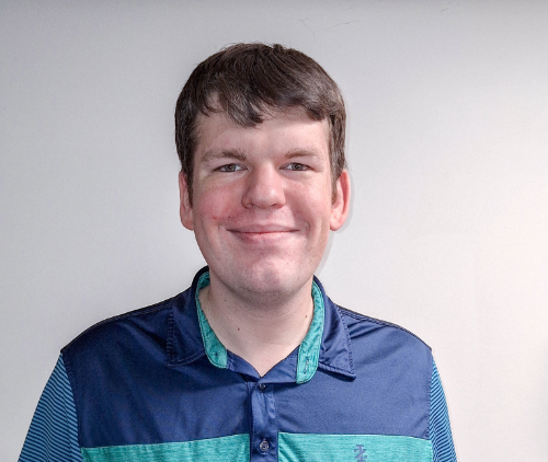 A person wearing a blue collared shirt smiles at the camera against a plain white background.