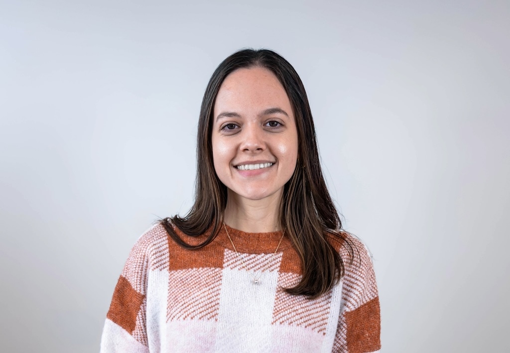 A person with long brown hair wearing a plaid sweater smiles against a plain background.