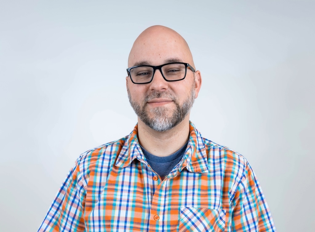 A person with a bald head and a beard is wearing glasses and a plaid shirt, standing against a plain white background.