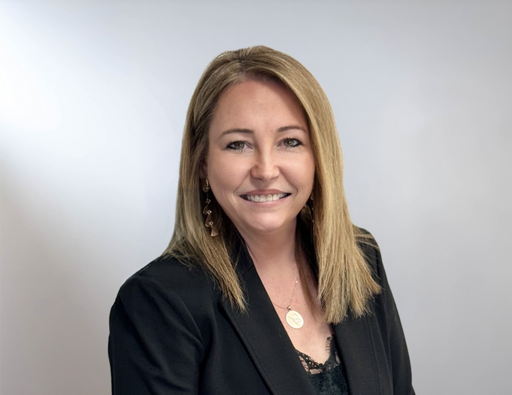 A woman with straight blonde hair, wearing a black blazer and a necklace, smiles at the camera against a plain light background.
