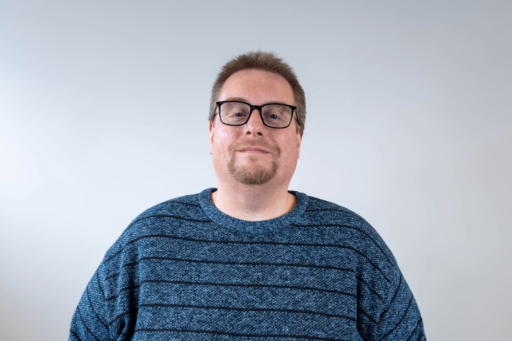 A person wearing glasses and a blue-striped sweater stands against a light gray background.