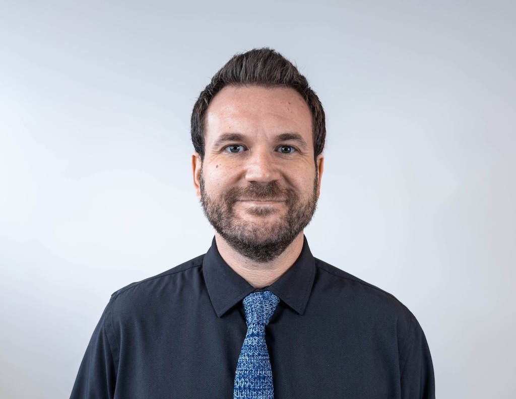 A man with a beard and short hair, wearing a dark shirt and patterned blue tie, smiles slightly against a light gray background.