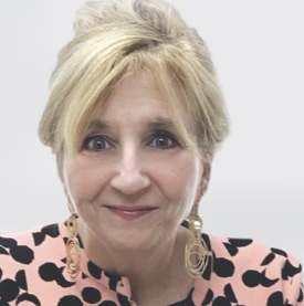 Woman with short blonde hair, wearing large gold earrings and a peach top with black polka dots, smiling at the camera against a plain light background.