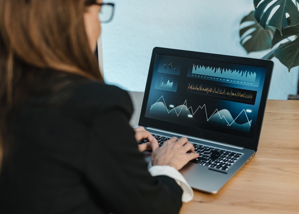 A woman in business attire sits at a desk, analyzing various data charts and graphs displayed on a laptop screen.