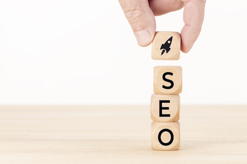 A hand places a wooden block with a rocket icon on top of three blocks stacked vertically spelling "SEO" on a light wood surface.