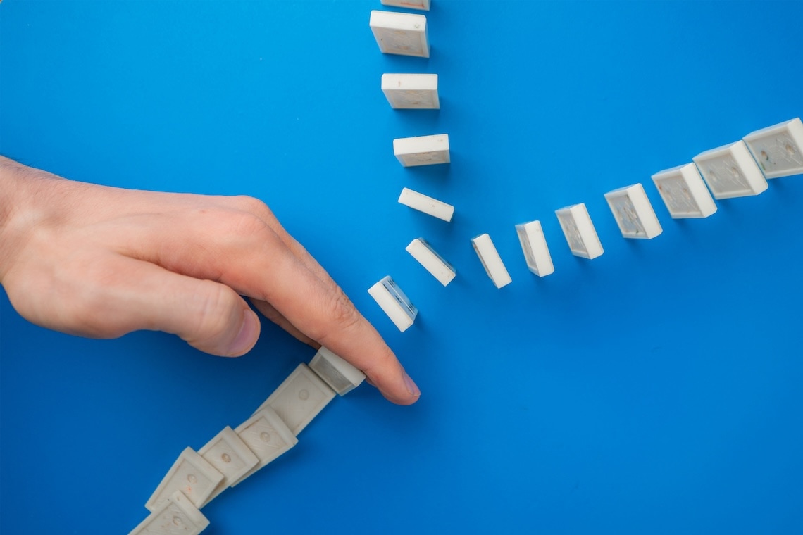 A hand stops the fall of white dominoes arranged in a Y-shape pattern on a blue surface.