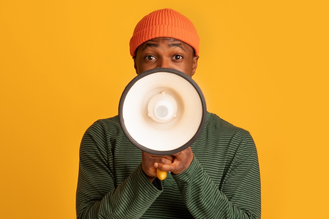 A person in a striped green shirt and orange beanie holds a megaphone to their mouth, standing in front of a solid yellow background.