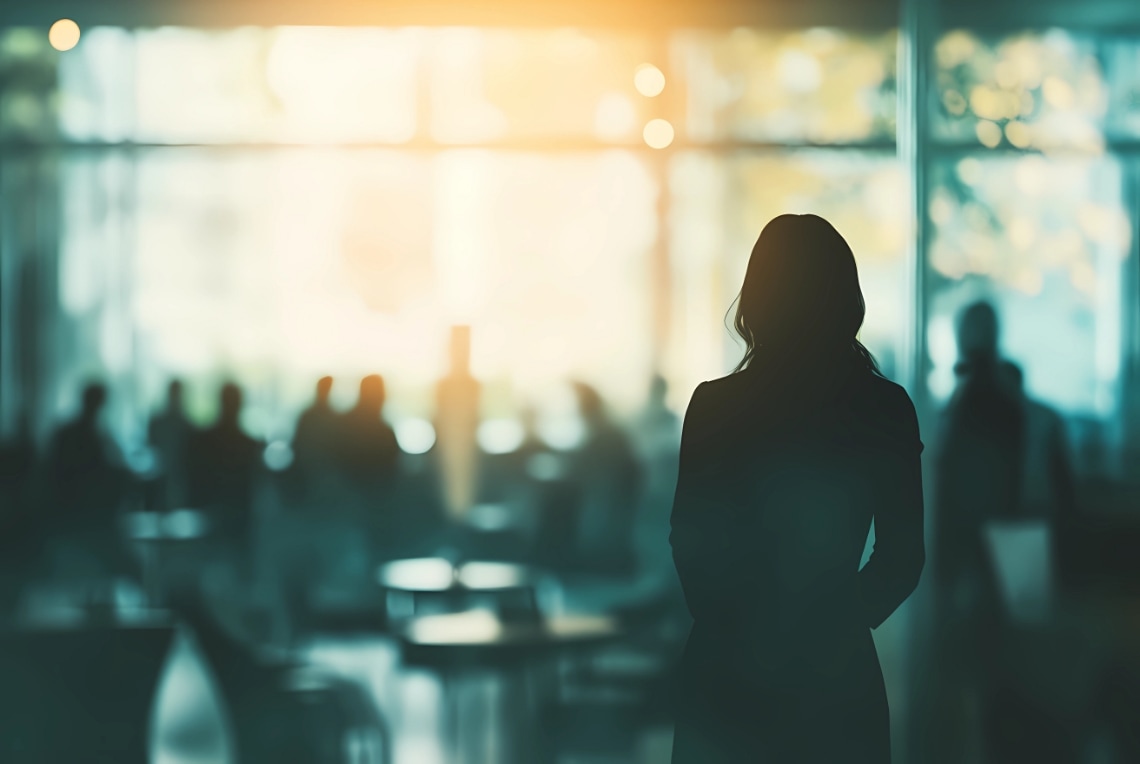 A silhouetted person stands indoors, embodying inclusive leadership as they face a sunlit area with several blurred figures seated at tables in the background.