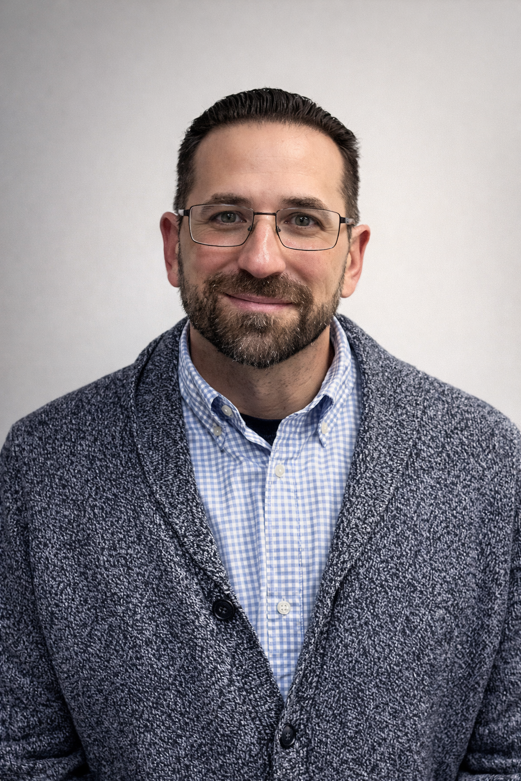 A man wearing glasses, a blue checkered shirt, and a grey textured cardigan stands in front of a plain light background, looking at the camera and smiling slightly.